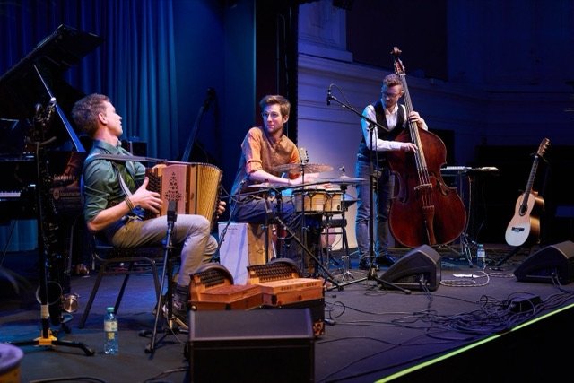 Sturm & Klang auf der Bühne des Lorely-Saals in Wien, in blauem Licht: von links Nik Jam an der Steirischen Harmonika, Jonny Kölbl an der Perkussion und Max Ganster am Kontrabass.