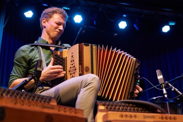 Nik Jam lacht beim Spielen der Steirischen Harmonika, im Hintergrund Bühnenscheinwerfer.