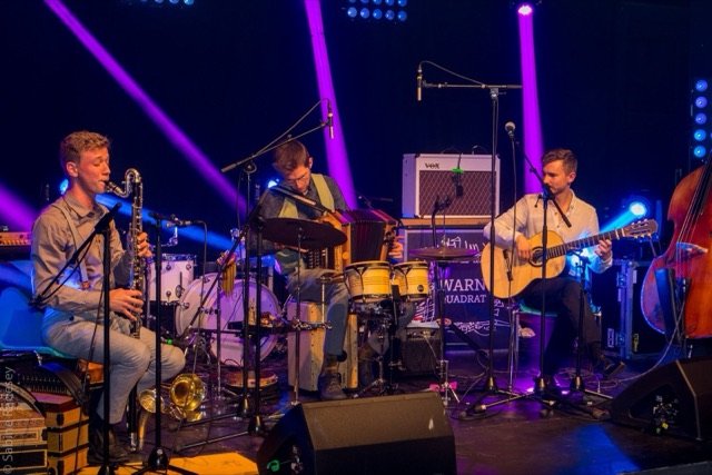 Sturm & Klang live in violettem Scheinwerferlicht: Nik Jam mit Bassklarinette, Jonny Kölbl am Schlagwerk und Max Ganster mit Gitarre.