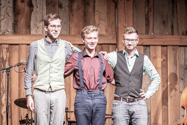 Nik Jam, Max Ganster und Jonny Kölbl Arm in Arm vor einer Holzwand, lachend nach dem Konzert.