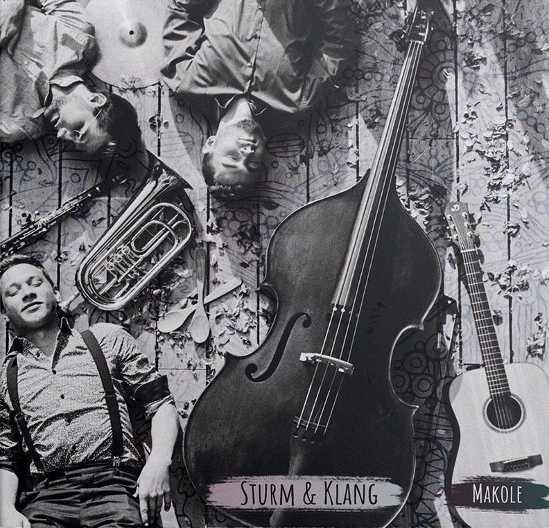 CD-Cover „Makole“: Die drei Musiker liegen in Schwarzweiß auf einem Holzboden zwischen Kontrabass, Gitarren, Harmonika und Blasinstrumenten.