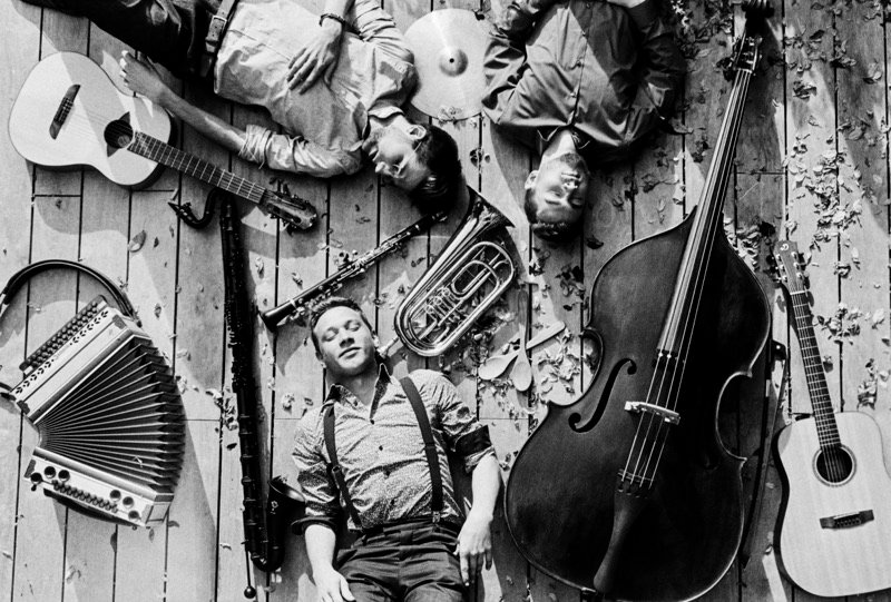 Schwarzweiß-Foto von oben: Die drei Musiker liegen zwischen ihren Instrumenten auf einem Holzboden.