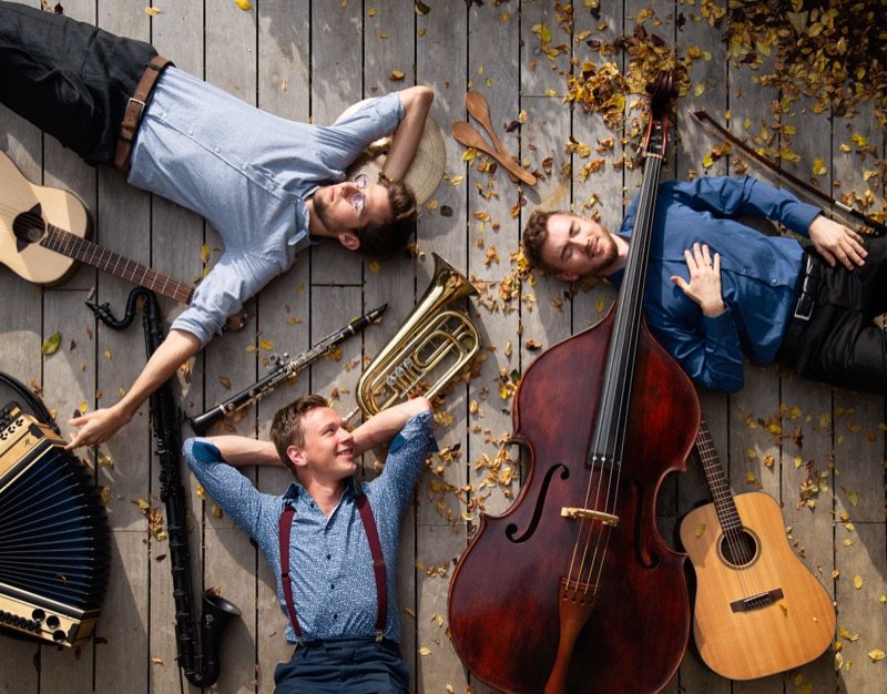 Farbfoto von oben: Die drei Musiker liegen lachend zwischen Herbstlaub, Kontrabass, Gitarren und Harmonika auf einem Holzboden.